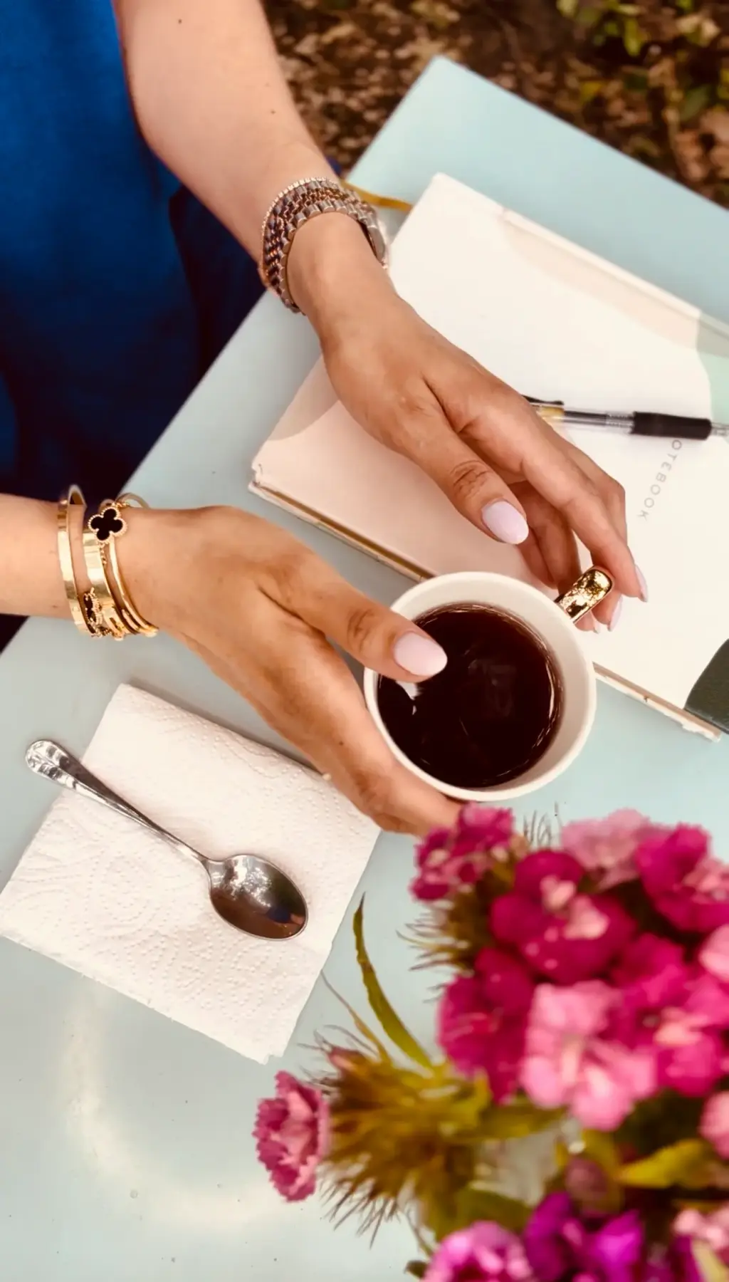 Hands holding a cup of tea on a table with flowers and a notebook.